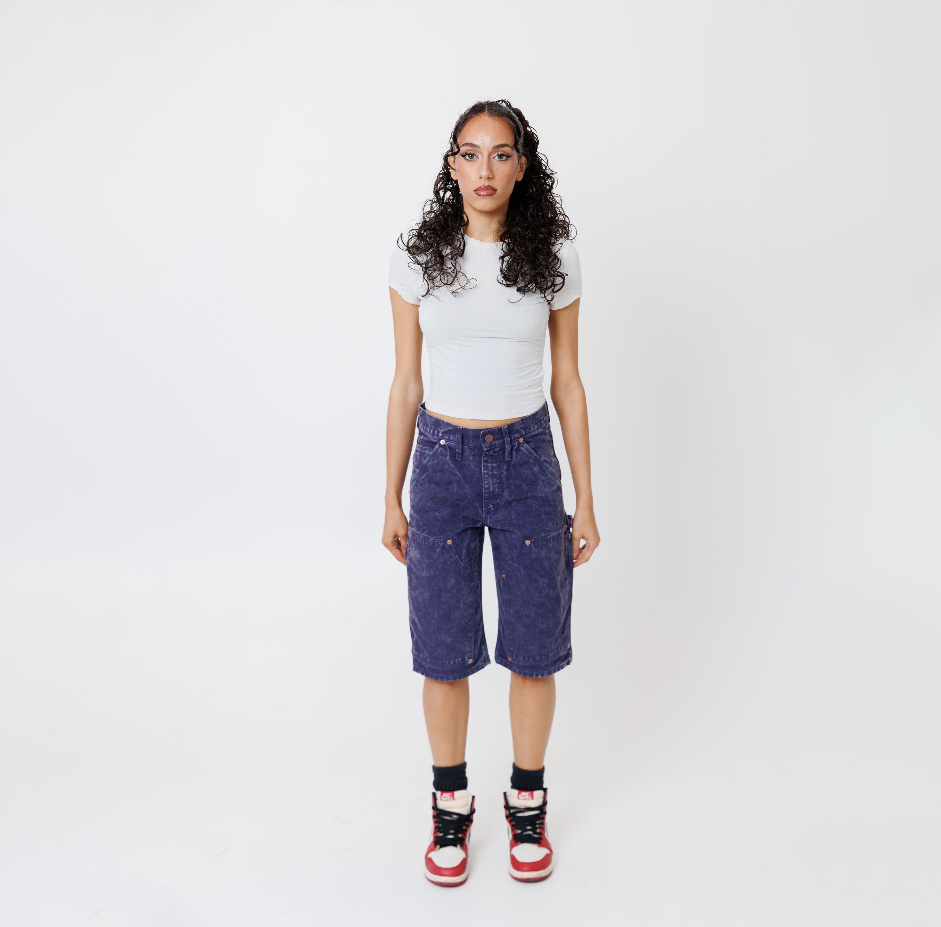 Female model wearing a white t-shirt and Wisdom of age canvas carpenter shorts in purple color on a light gray background