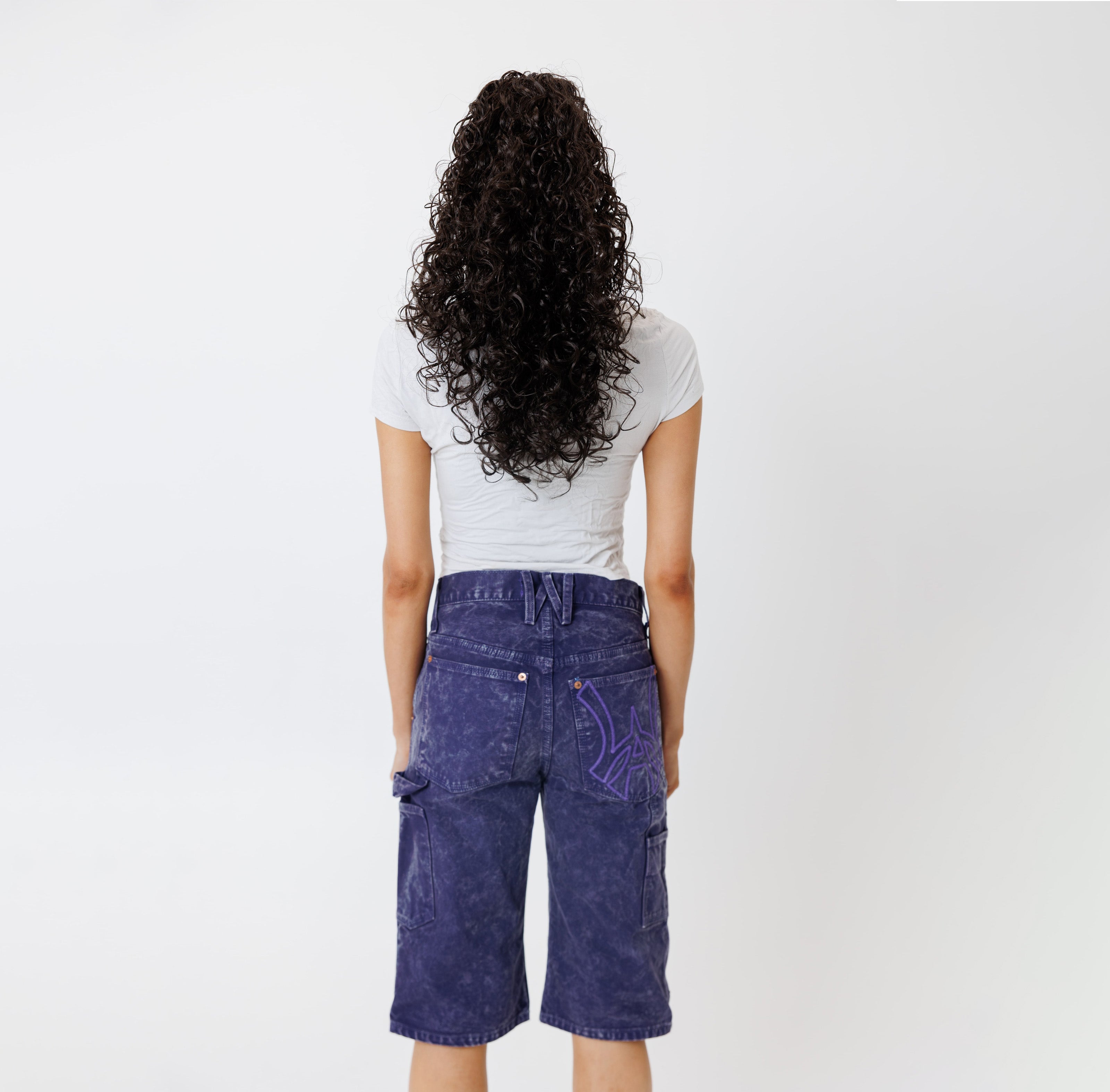 Rear view of a Female model wearing a white t-shirt and Wisdom of age canvas carpenter shorts in purple color on a light gray background