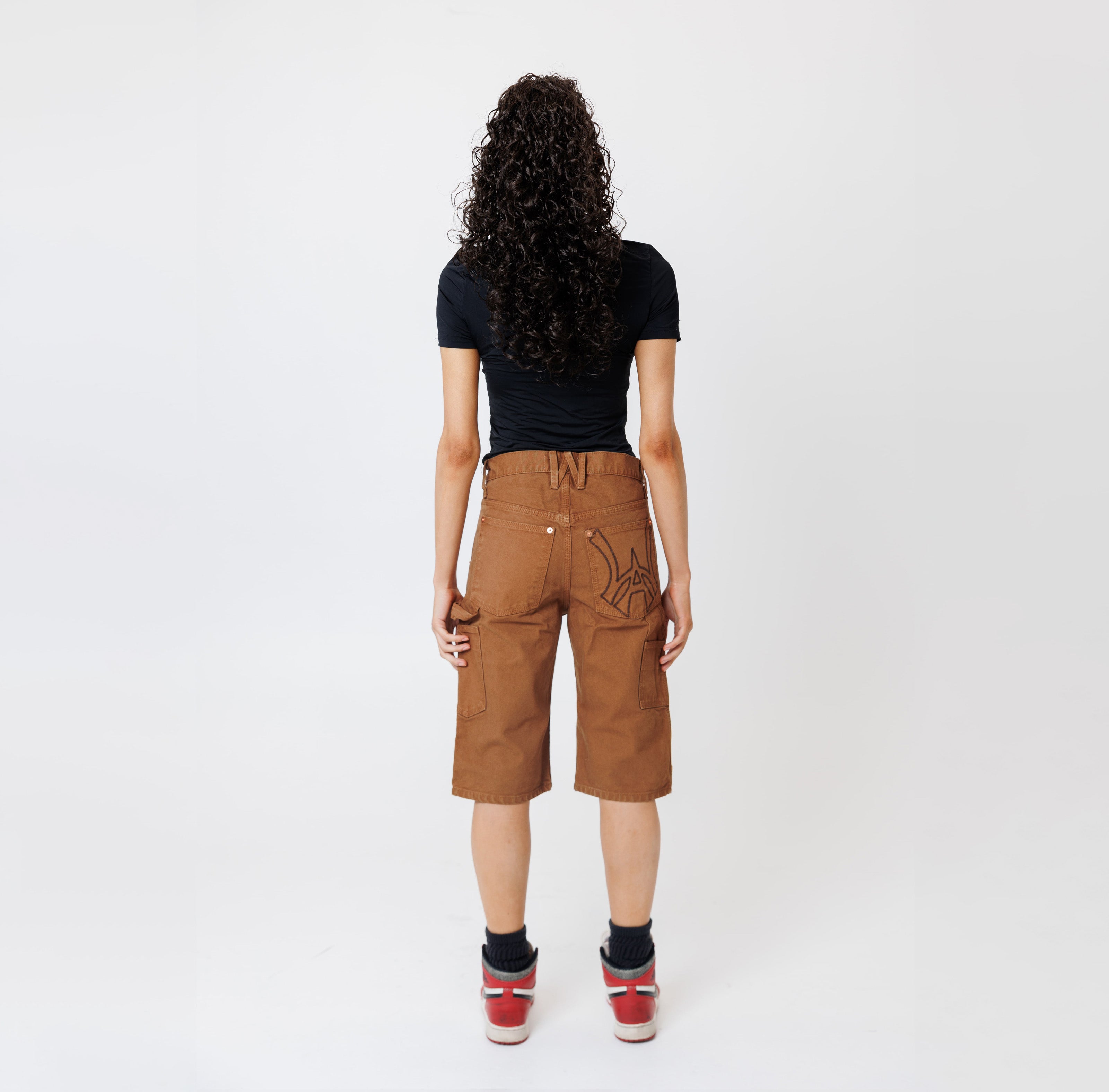 Rear view of a Female model wearing a black t-shirt and Wisdom of age canvas carpenter shorts in brick color on a light gray background