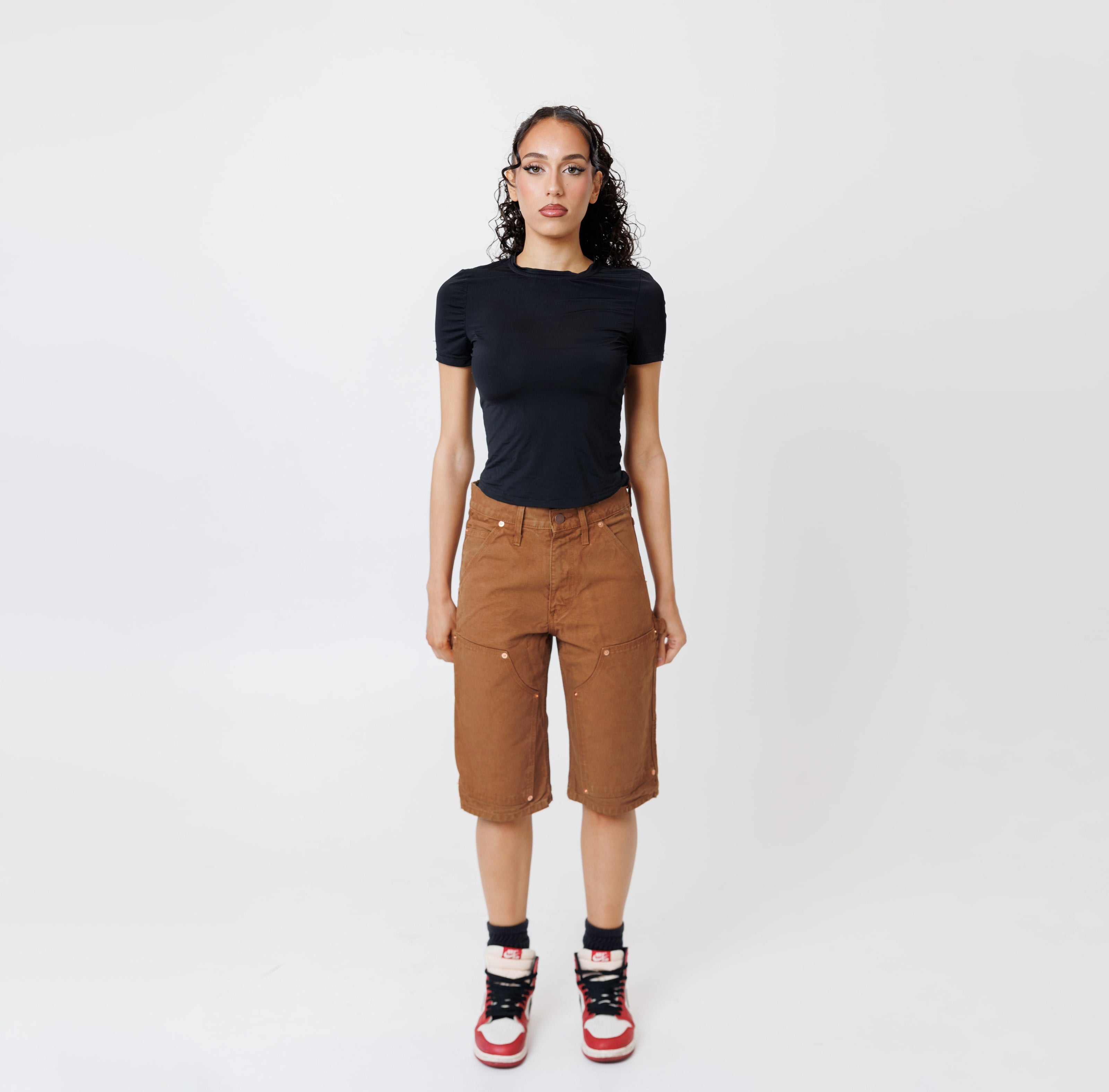 Female model wearing a black t-shirt and Wisdom of age canvas carpenter shorts in brick color on a light gray background