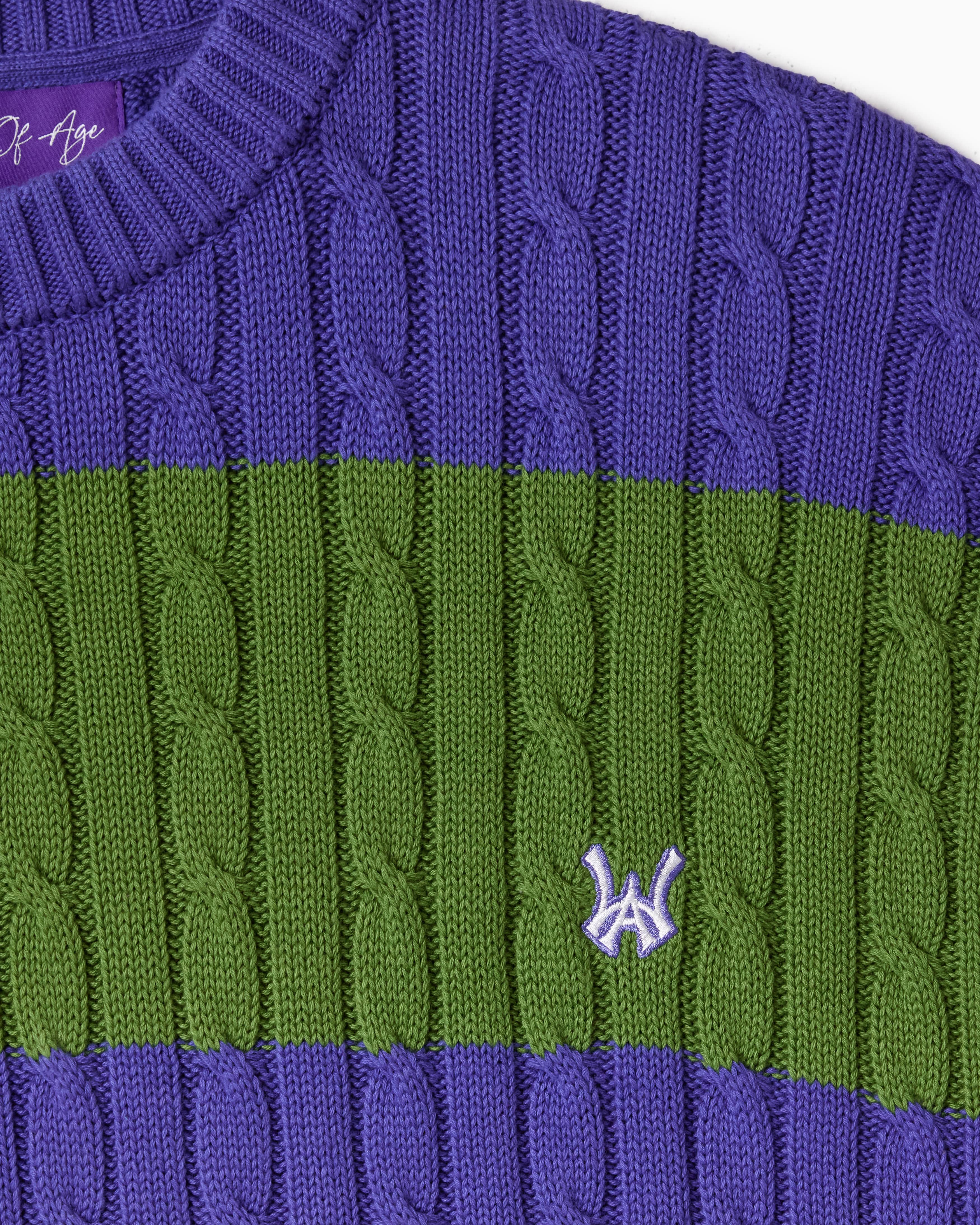 Close-up of a purple and green cable knit T-Shirt with WA logo on a white background.