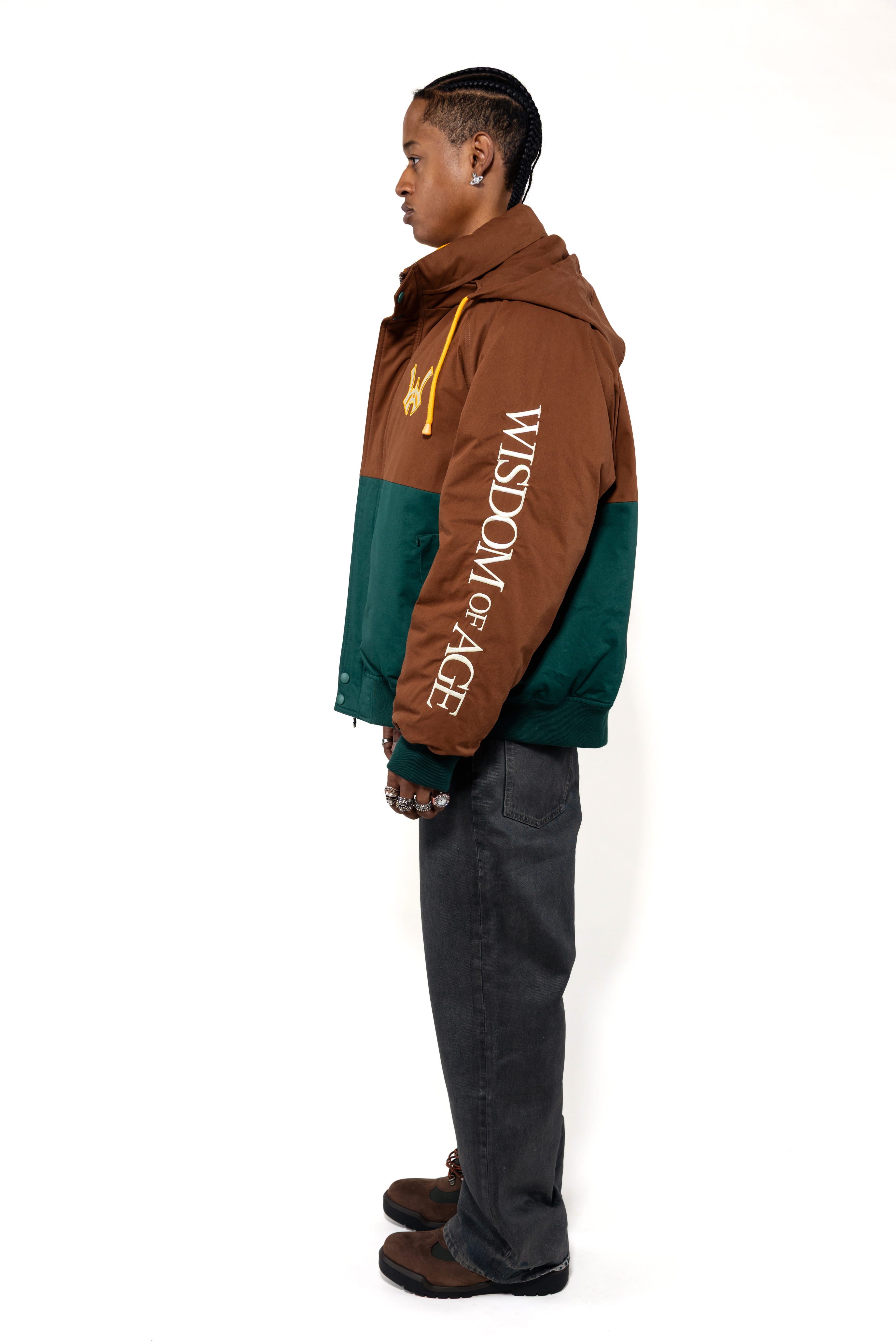 Side view of a Male Model wearing a brown and forest green jacket with yellow accents on a white background. 'Wisdom Of Age' written on the sleeve.