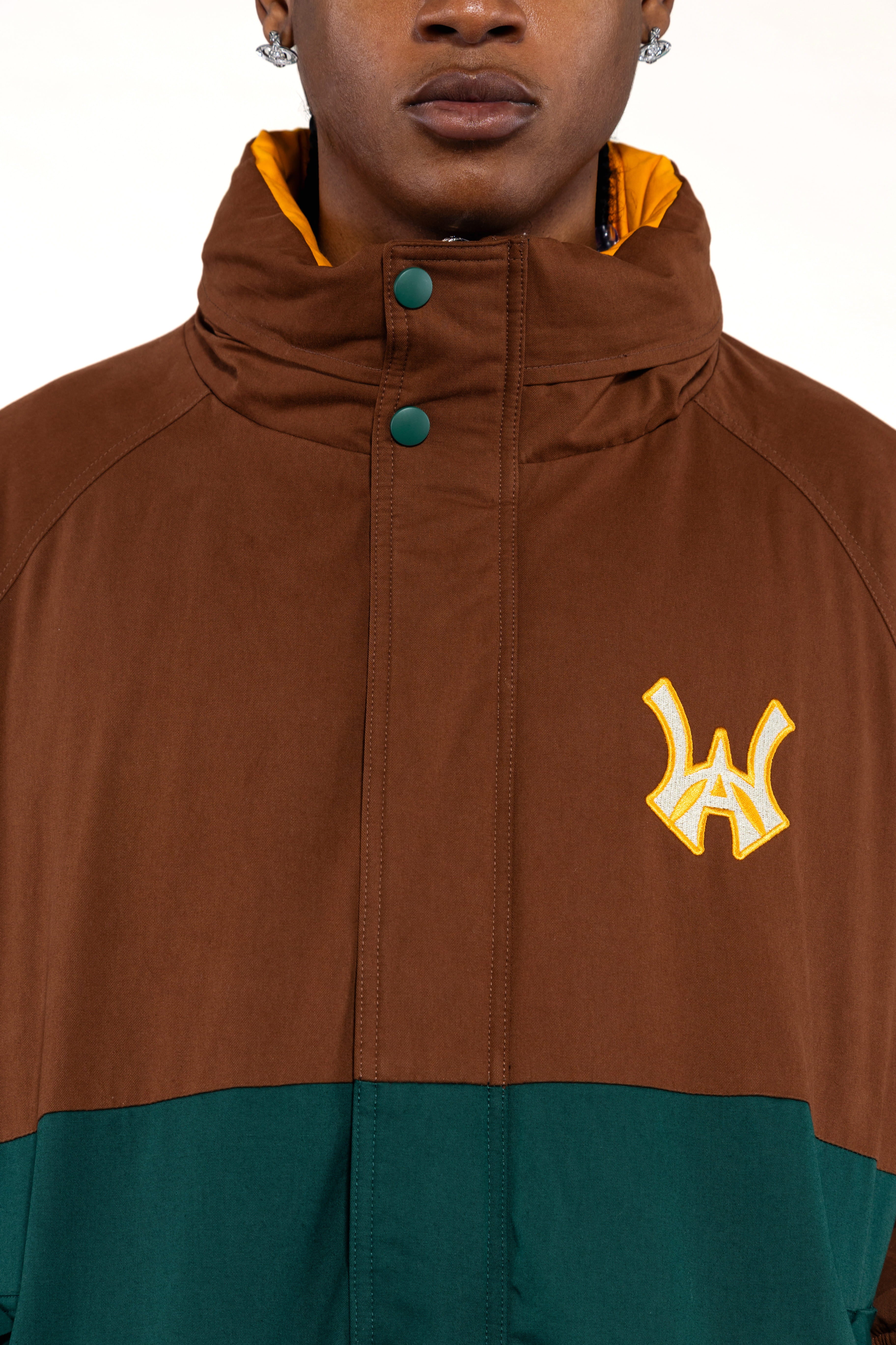 Close up view of a Male Model wearing a brown and forest green jacket with yellow accents on a white background. 'WA' Logo embroidered on the left chest.