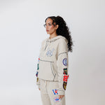 Female model wearing a theW.O.A. Rugby Club Sweat suit in Sandstone with multiple embroidered patches on the sleeves and pant legs on a white background