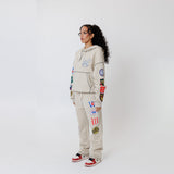Female model wearing a theW.O.A. Rugby Club Sweat suit in Sandstone with multiple embroidered patches on the sleeves and pant legs on a white background
