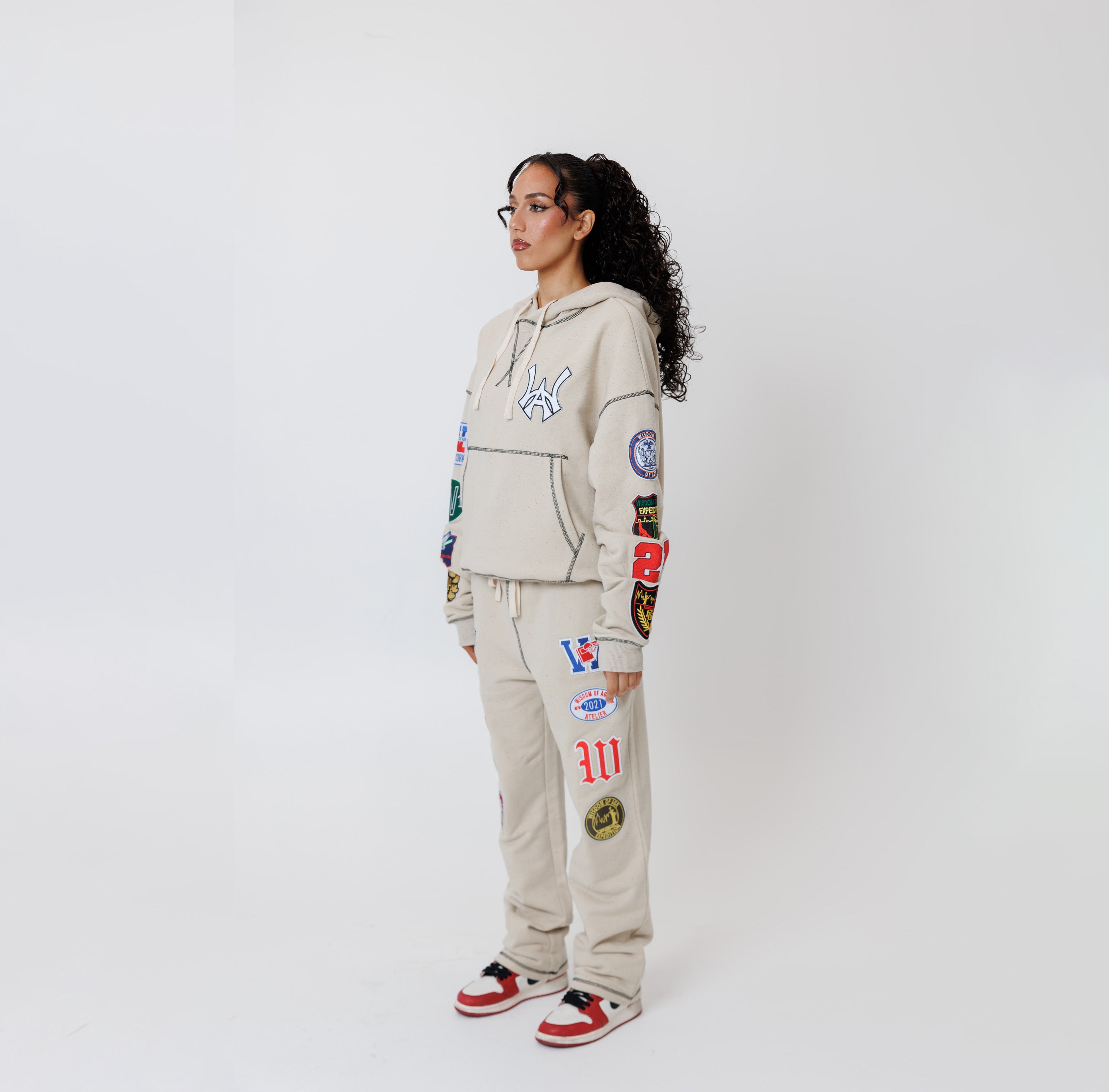 Female model wearing a theW.O.A. Rugby Club Sweat suit in Sandstone with multiple embroidered patches on the sleeves and pant legs on a white background