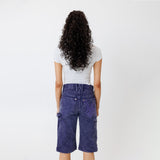Rear view of a Female model wearing a white t-shirt and Wisdom of age canvas carpenter shorts in purple color on a light gray background