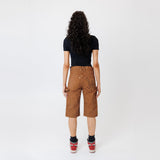 Rear view of a Female model wearing a black t-shirt and Wisdom of age canvas carpenter shorts in brick color on a light gray background