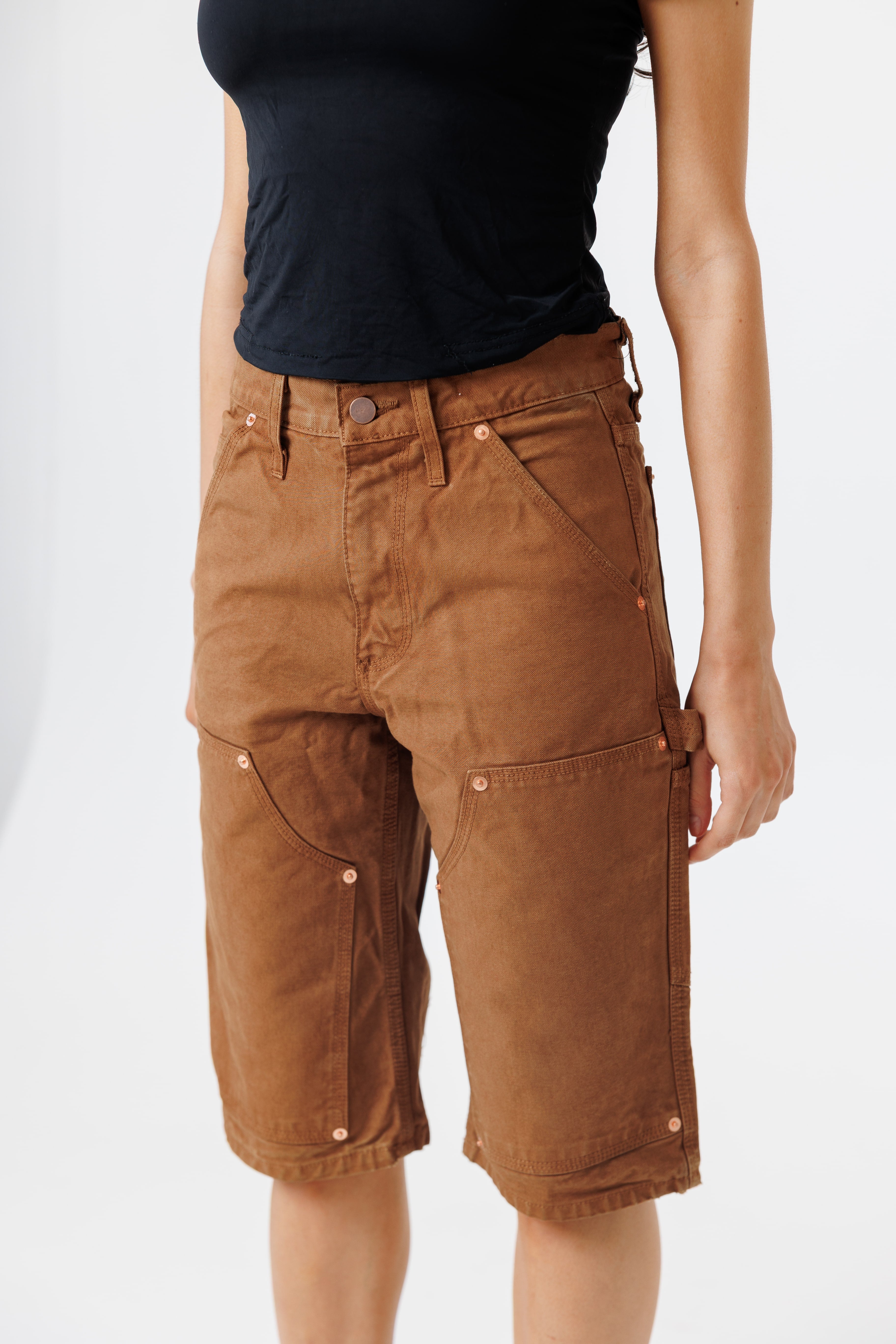 close up view of a Female model wearing a black t-shirt and Wisdom of age canvas carpenter shorts in brick color on a light gray background