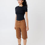 Side view of a Female model wearing a black t-shirt and Wisdom of age canvas carpenter shorts in brick color on a light gray background