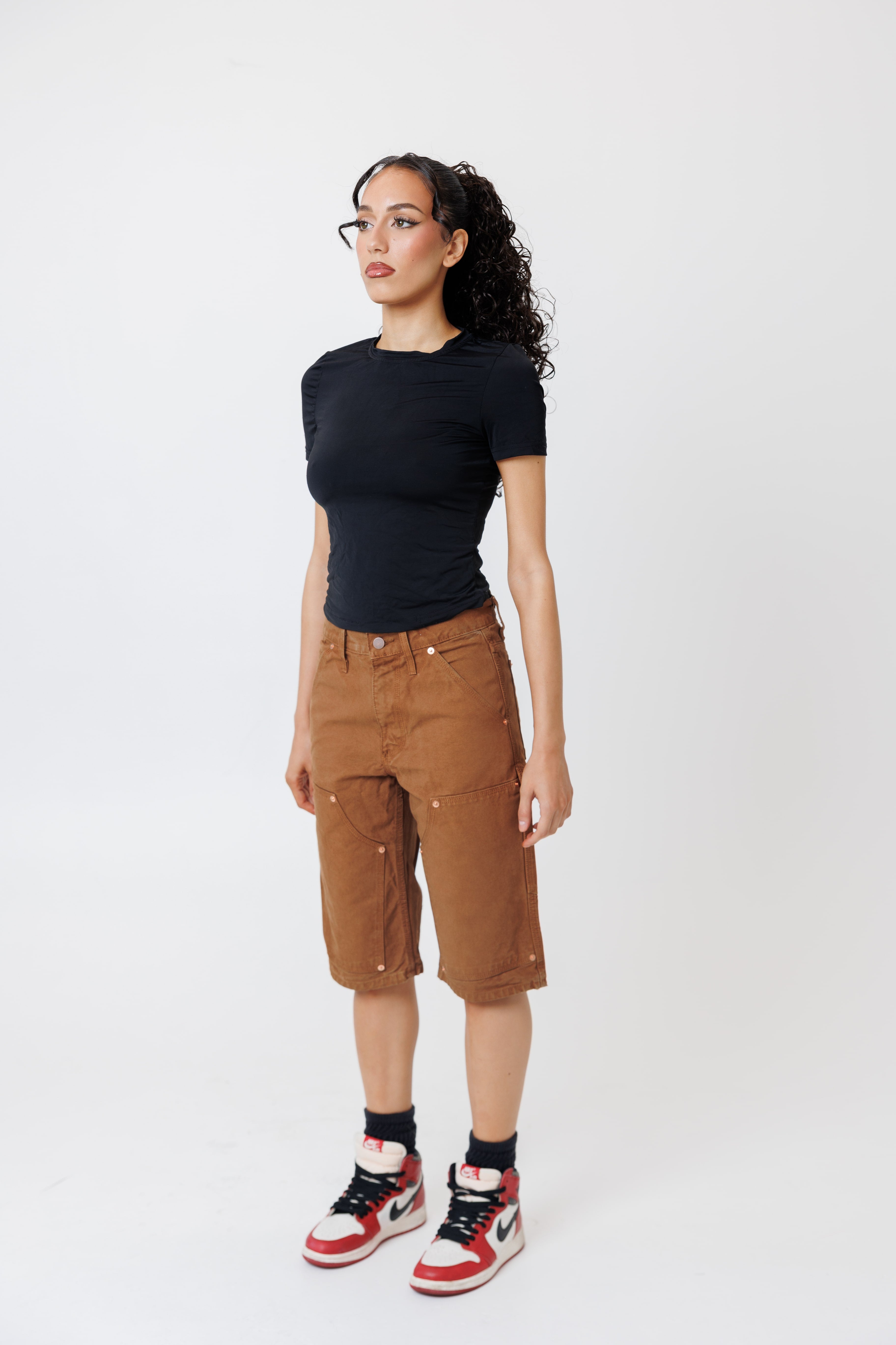 Side view of a Female model wearing a black t-shirt and Wisdom of age canvas carpenter shorts in brick color on a light gray background