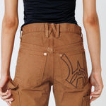Rear close up view of a Female model wearing a black t-shirt and Wisdom of age canvas carpenter shorts in brick color on a light gray background
