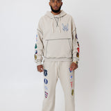 male model wearing a the W.O.A. Rugby Club Sweat suit in Sandstone with multiple embroidered patches on the sleeves and pant legs on a white background