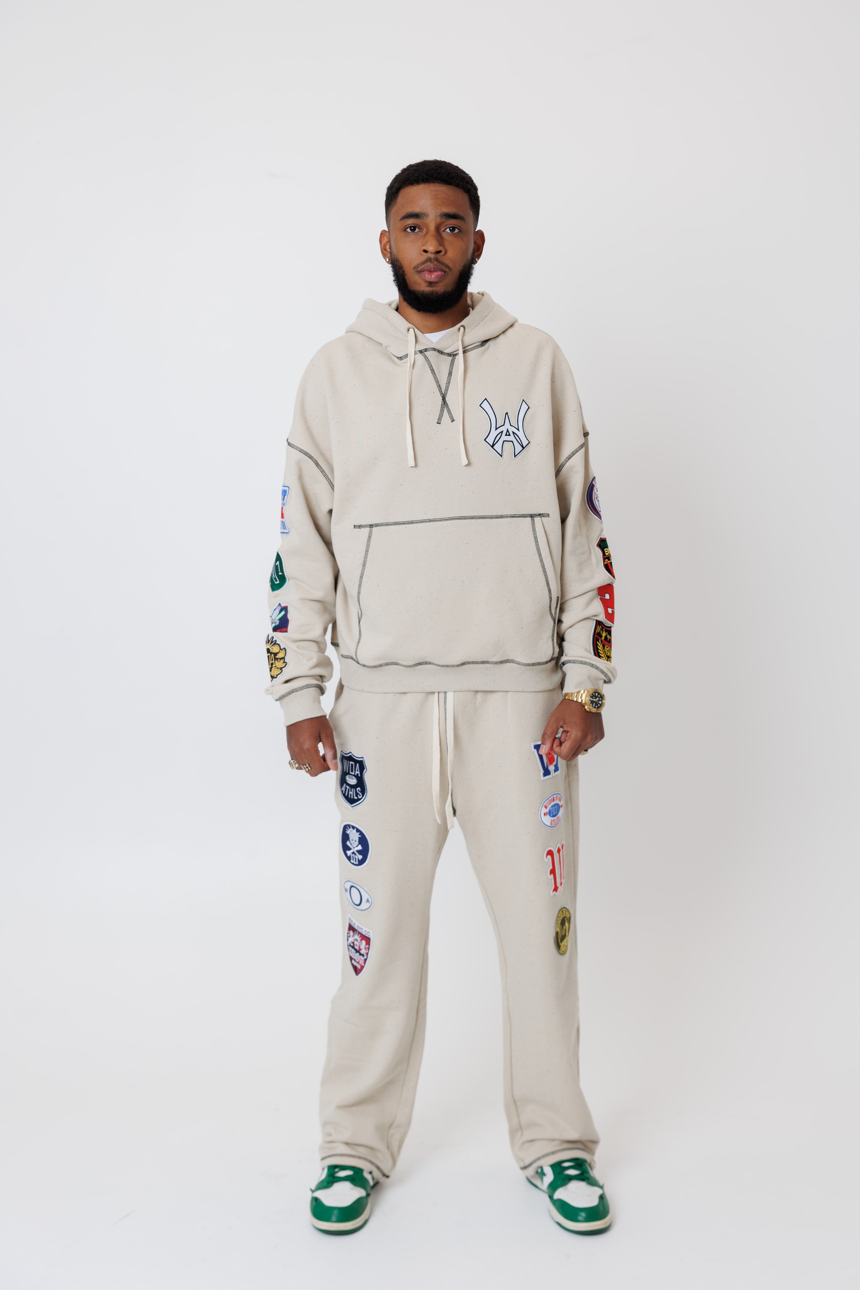 male model wearing a the W.O.A. Rugby Club Sweat suit in Sandstone with multiple embroidered patches on the sleeves and pant legs on a white background