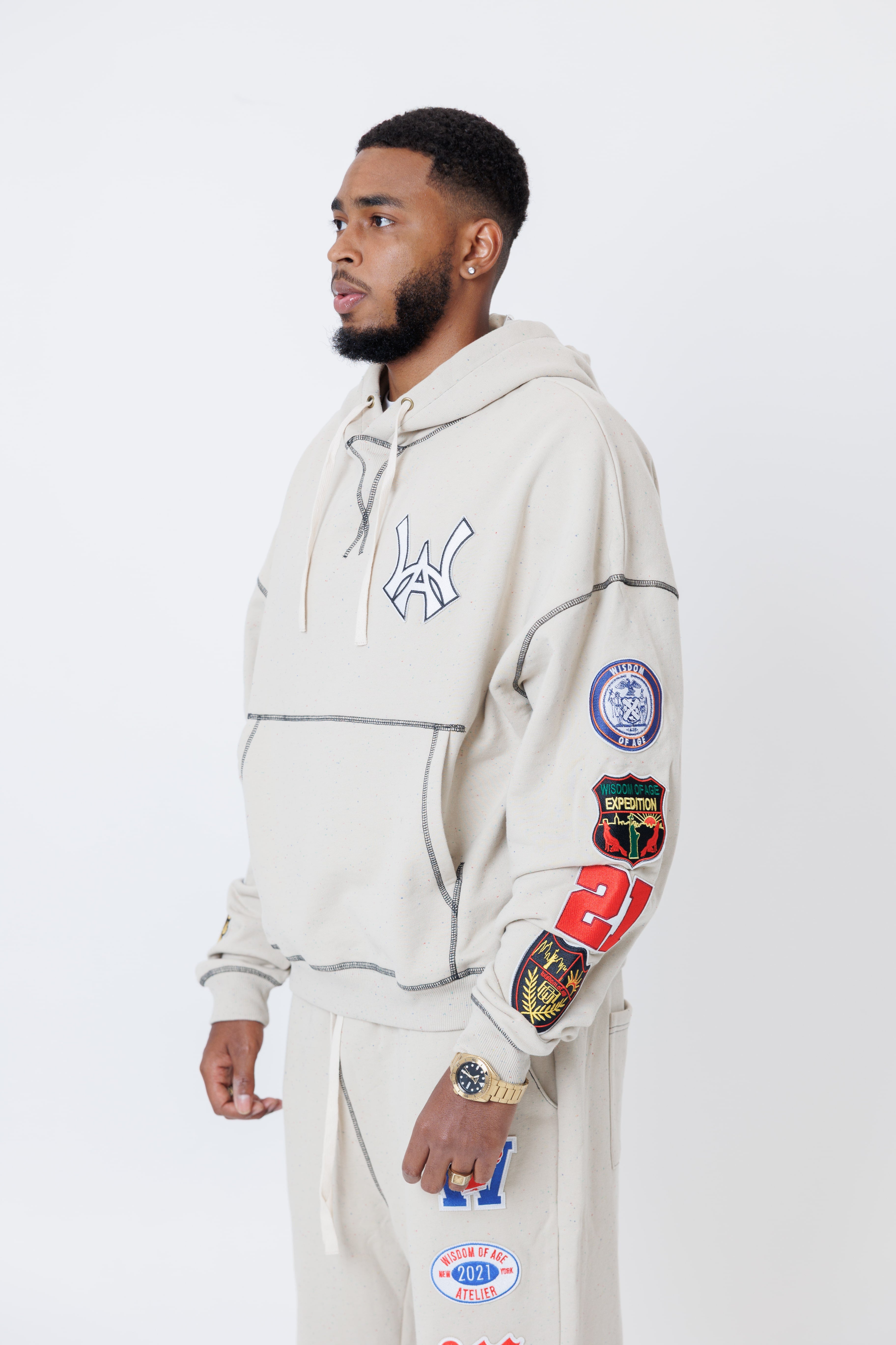 close up of a male model wearing a the W.O.A. Rugby Club Sweat suit in Sandstone with multiple embroidered patches on the sleeves and pant legs on a white background