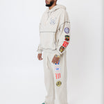 male model wearing a the W.O.A. Rugby Club Sweat suit in Sandstone with multiple embroidered patches on the sleeves and pant legs on a white background