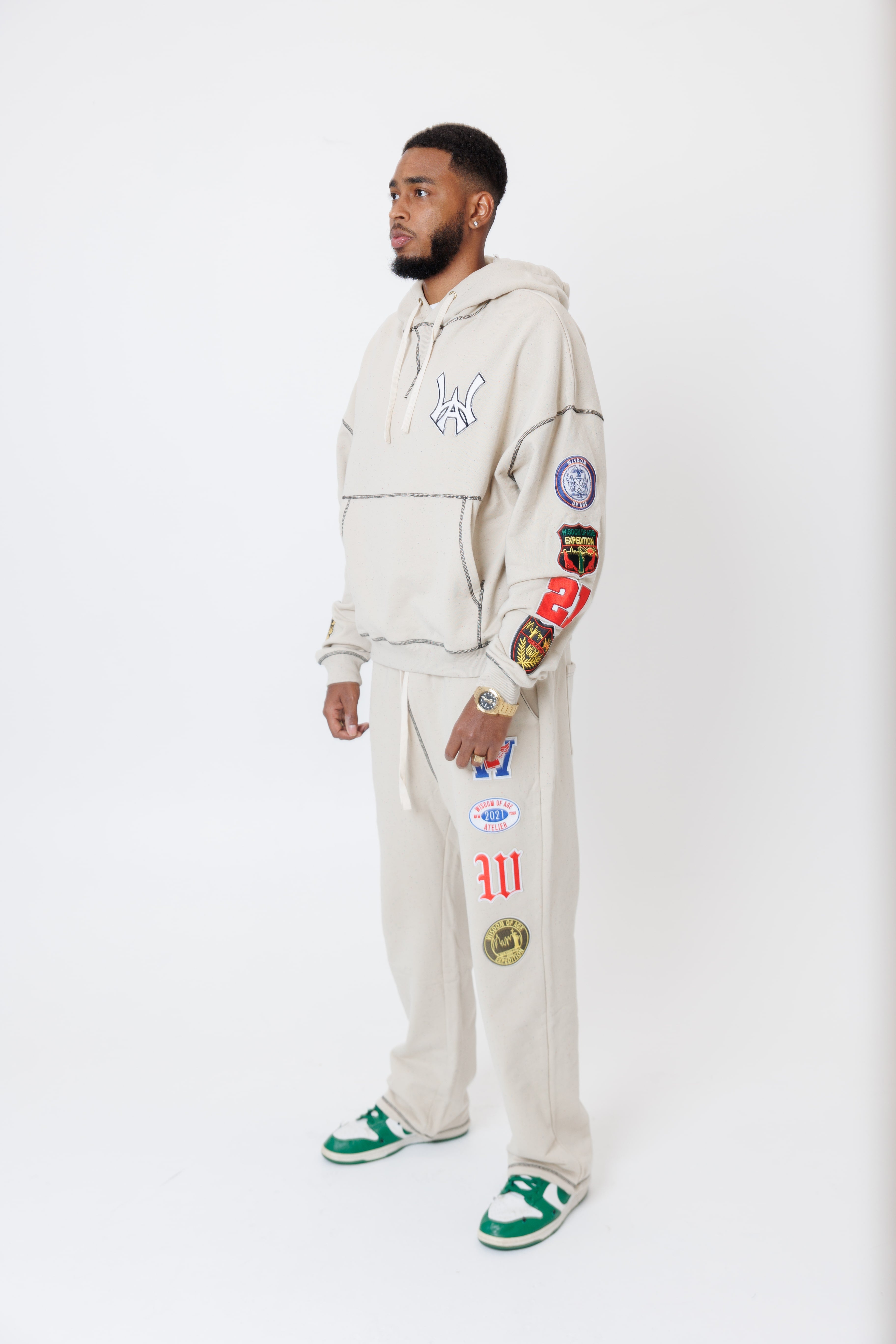 male model wearing a the W.O.A. Rugby Club Sweat suit in Sandstone with multiple embroidered patches on the sleeves and pant legs on a white background
