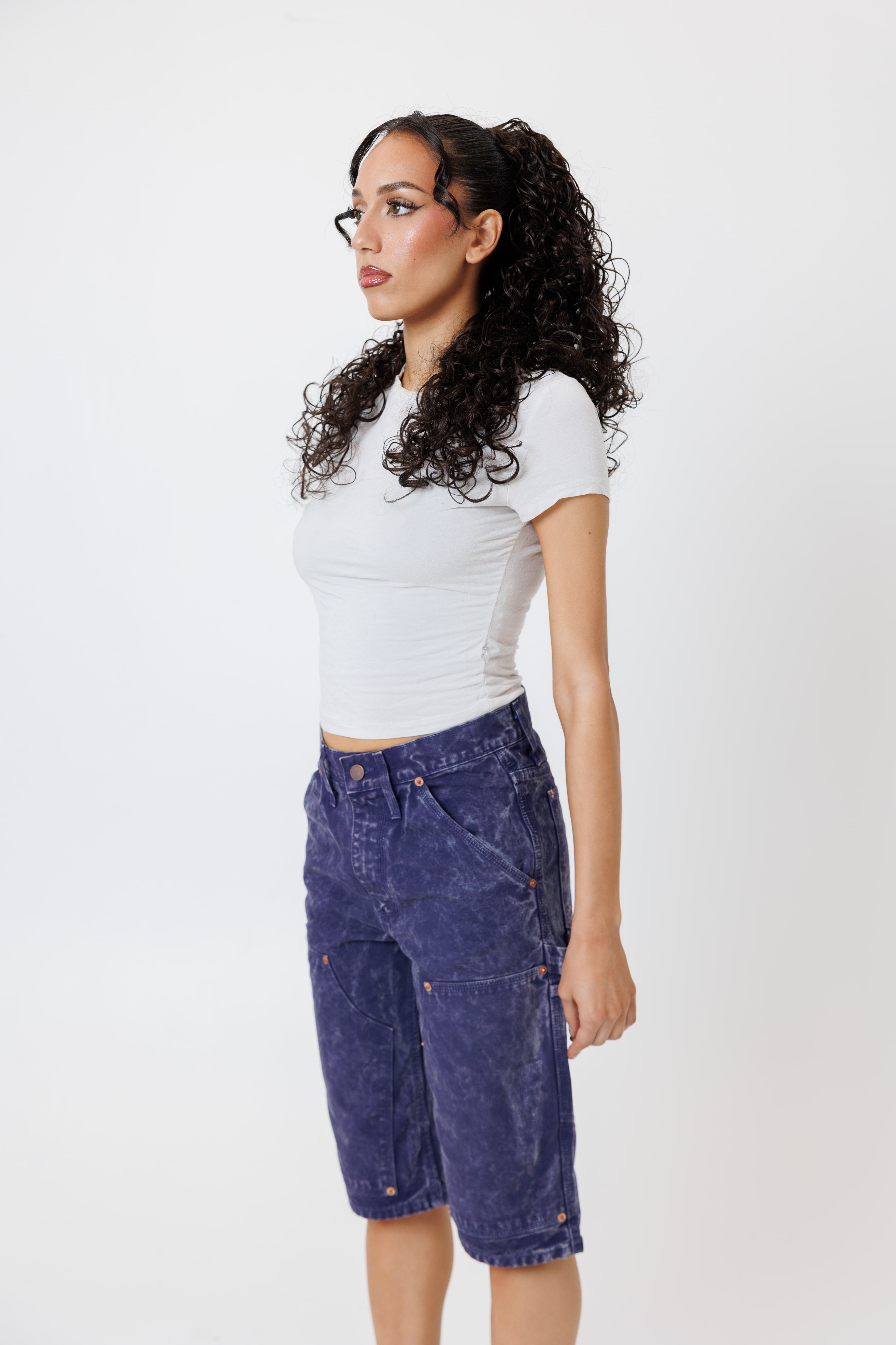 side view of a Female model wearing a white t-shirt and Wisdom of age canvas carpenter shorts in purple color on a light gray background
