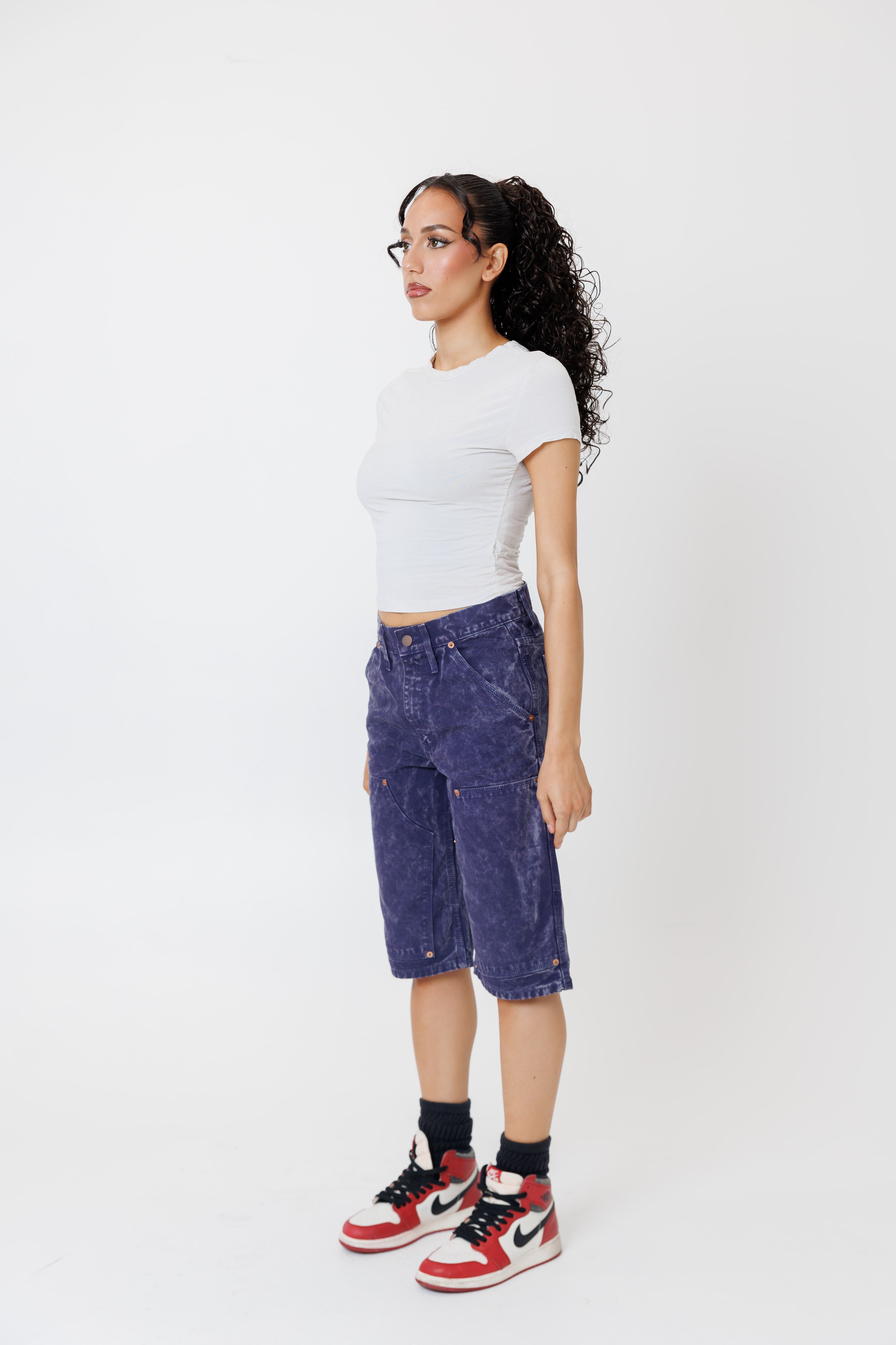 Side view of a Female model wearing a white t-shirt and Wisdom of age canvas carpenter shorts in purple color on a light gray background