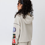 rear view of a Female model wearing a theW.O.A. Rugby Club Sweat suit in Sandstone with multiple embroidered patches on the sleeves and pant legs on a white background