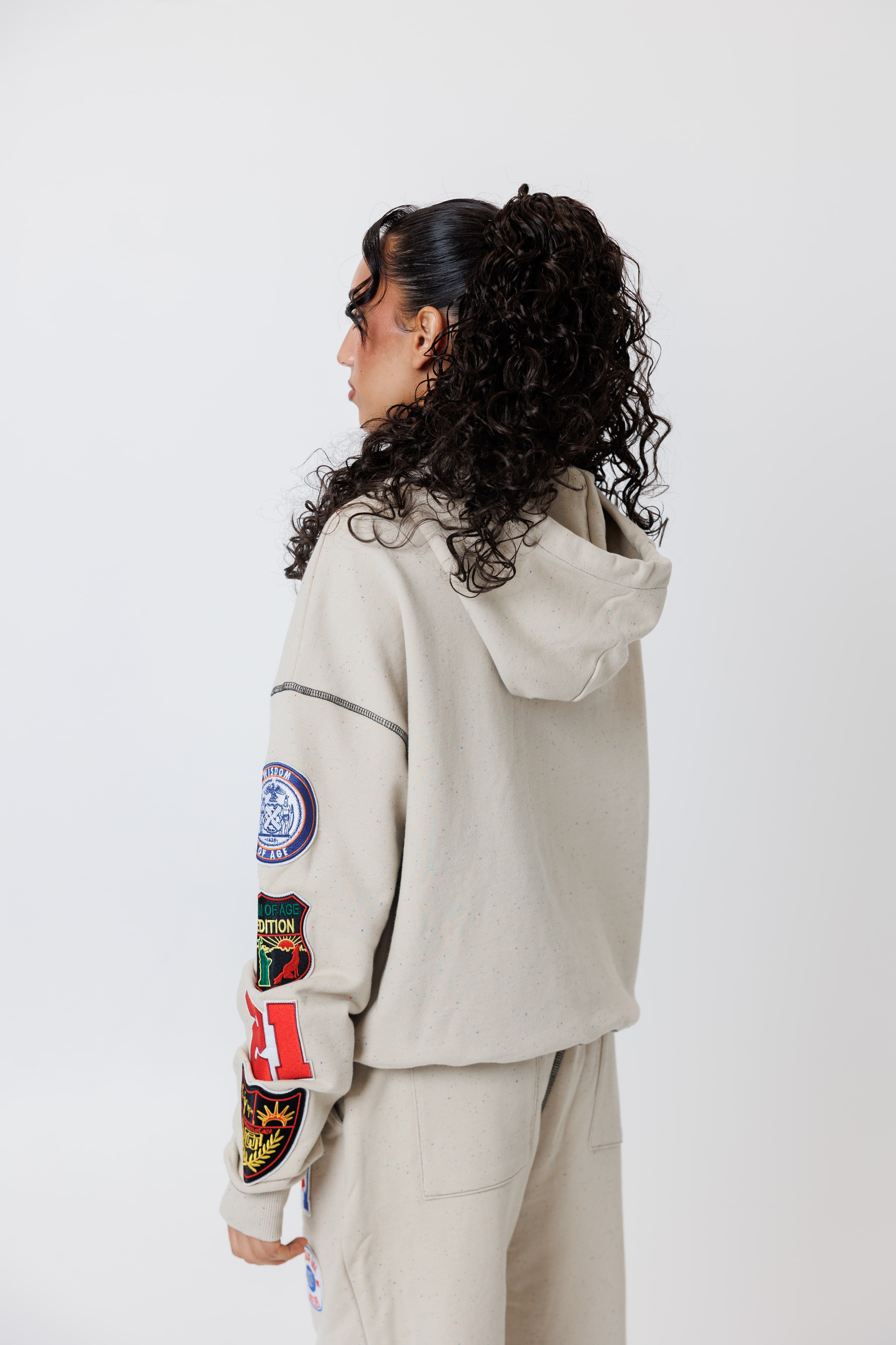 rear view of a Female model wearing a theW.O.A. Rugby Club Sweat suit in Sandstone with multiple embroidered patches on the sleeves and pant legs on a white background