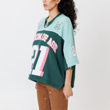side view of a female Model wearing a the WOA Football jersey V2 in the Mint color with 'Wisdom Of Age' and number '21' embroidered on the front. Model is wearing the WOA canvas carpenter shorts on a white background.