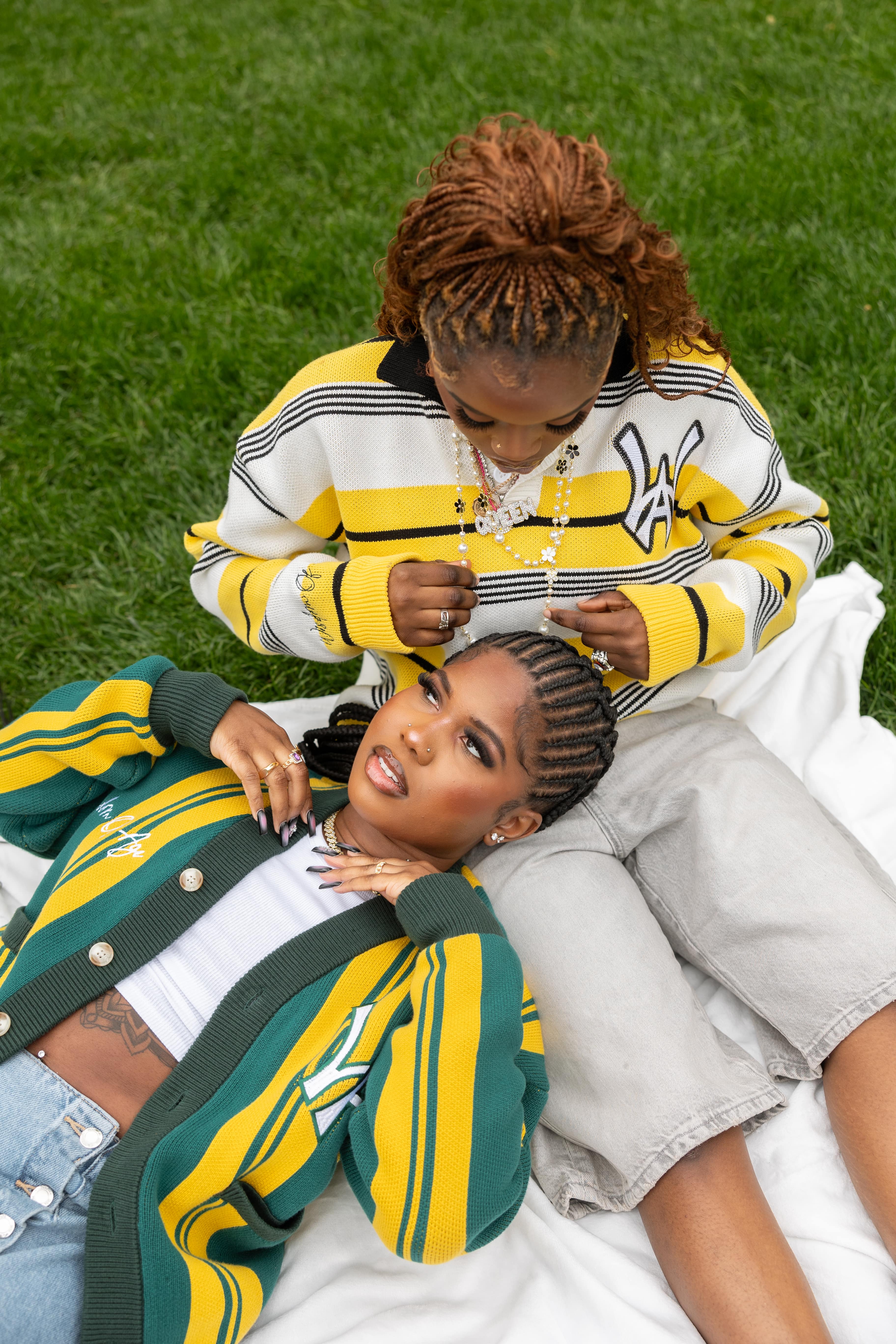 two women laying in a park. the woman on the right is wearing the WOA Rugby in black and yellow. the woman on the left, is wearing the WOA city cardigan in teal and yellow