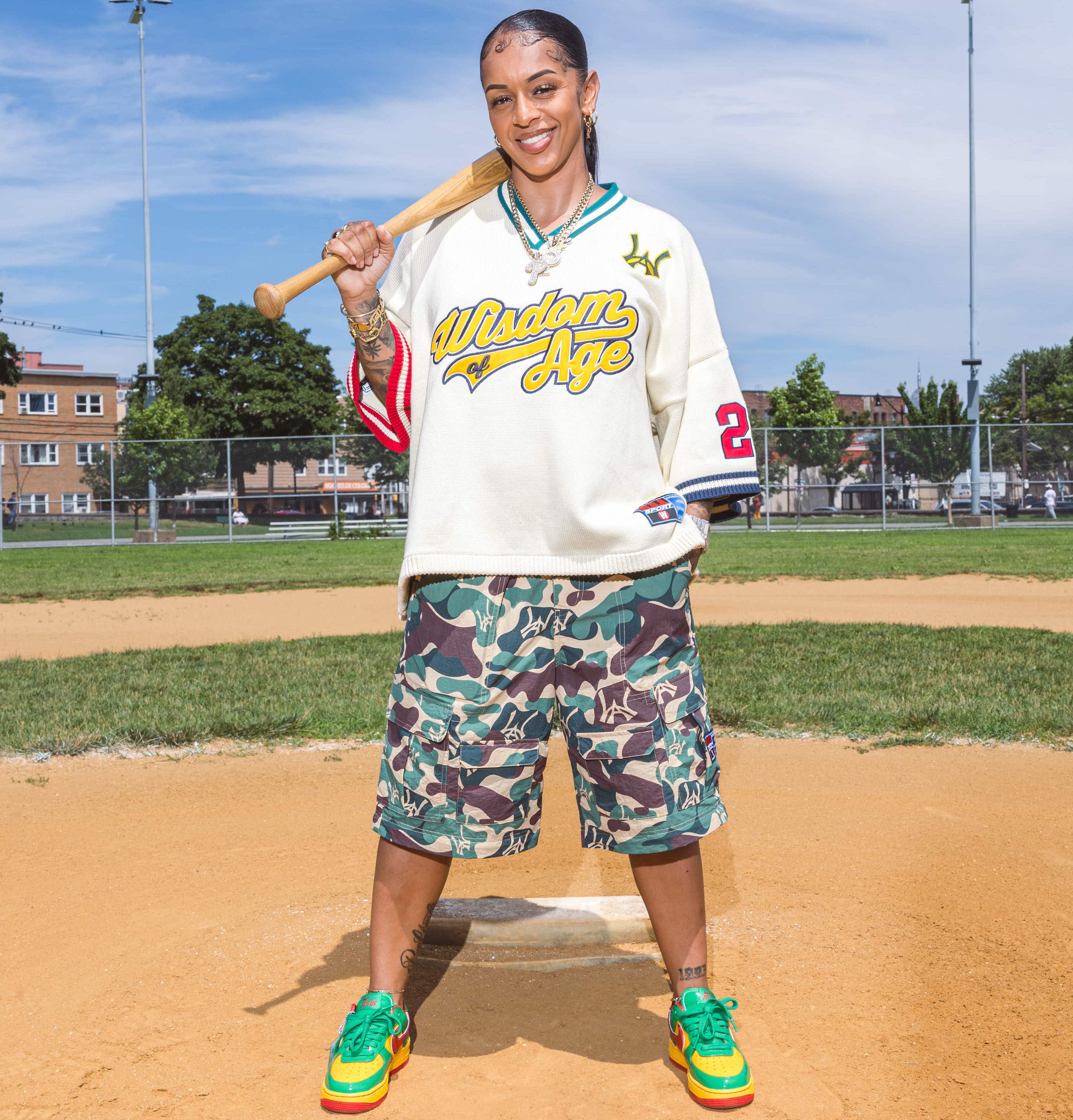 female model holding a baseball bat on a baseball field wearing the 'Wisdom of Age' knitted baseball jersey and Wisdom Of Age camouflage shorts.