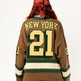 Rear view of a Female Model wearing the WOA Knitted Hockey Jersey with Embroidered 'NEW YORK' and the number 21 standing against a white background.