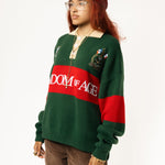 Side-front view of a Female Model wearing a green and red, knitted rugby sweater with 'Wisdom of Age' text on a white background.