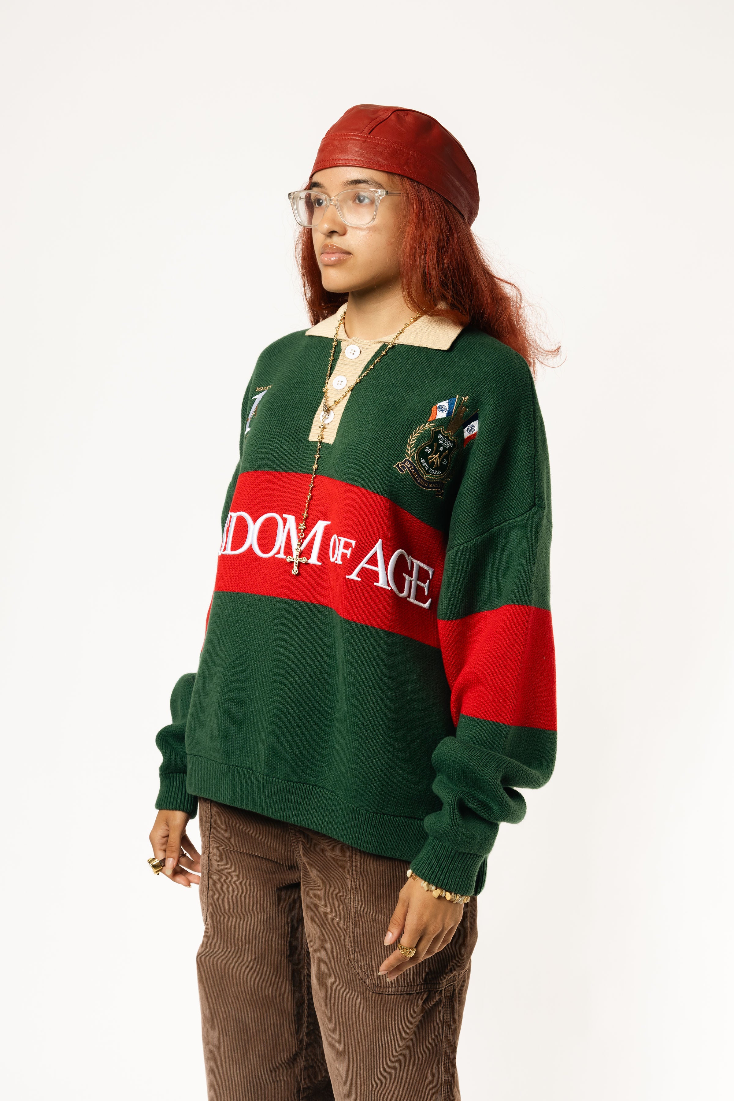 Side-front view of a Female Model wearing a green and red, knitted rugby sweater with 'Wisdom of Age' text on a white background.