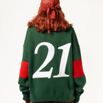 Rear view of a female Model wearing a green and red, knitted rugby sweater with '21' on a white background.