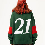 Rear view of a female Model wearing a green and red, knitted rugby sweater with '21' on a white background.