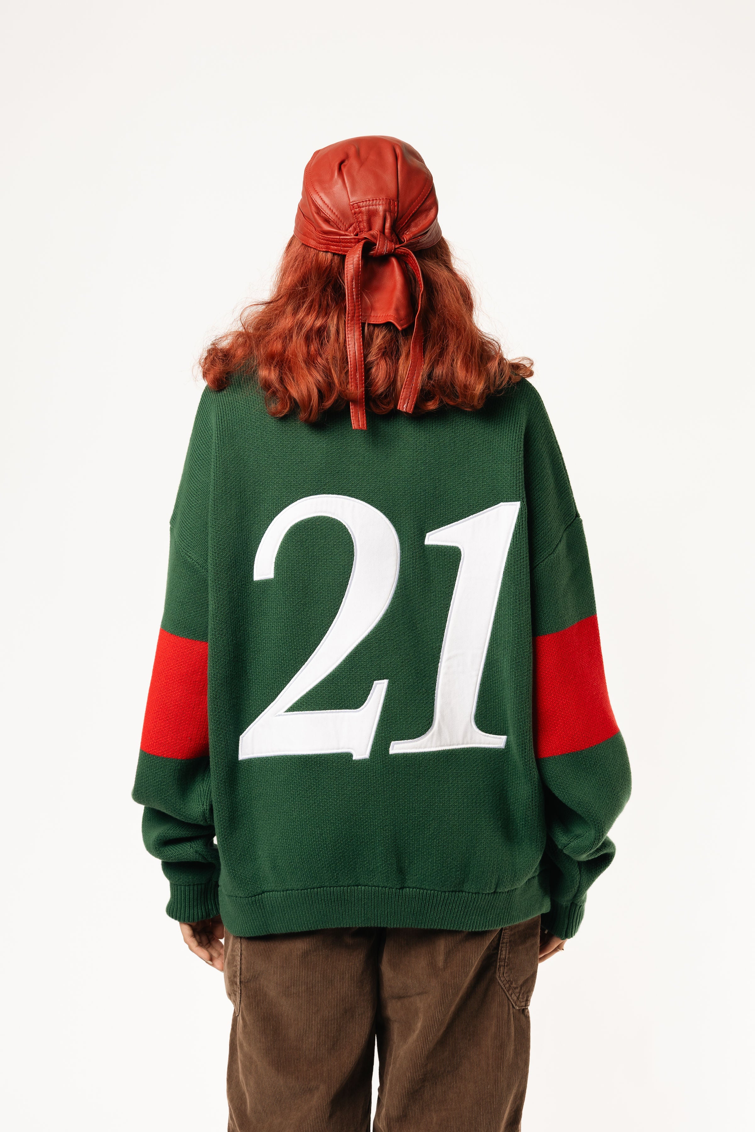 Rear view of a female Model wearing a green and red, knitted rugby sweater with '21' on a white background.