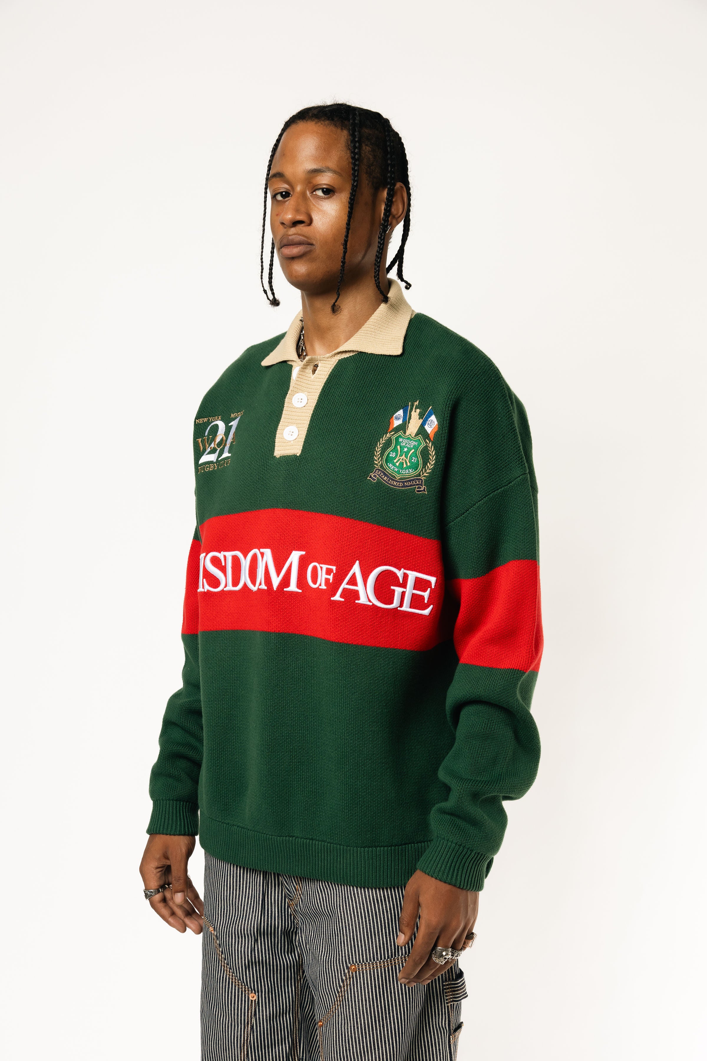 Front-side view of a Male Model wearing a green and red, knitted rugby sweater with 'Wisdom of Age' text on a white background.