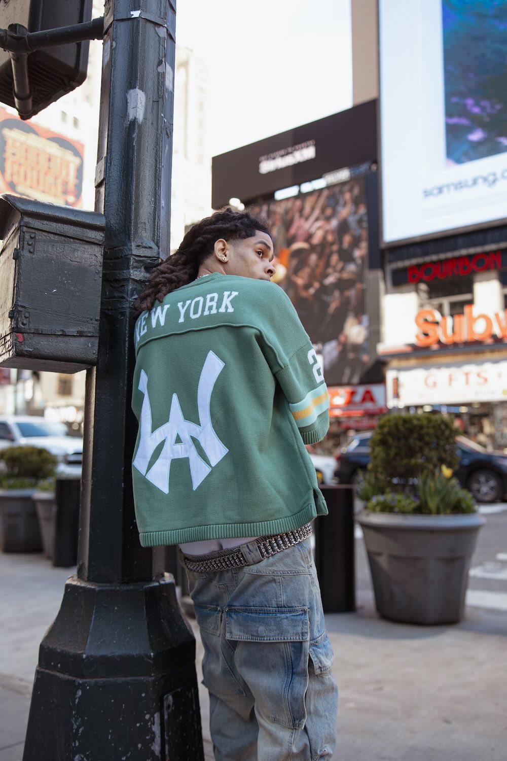 A male model leaning against a city street pole wearing a green knit jersey with New York spelled on the back and Wisdom of Age logo embroidered in white.
