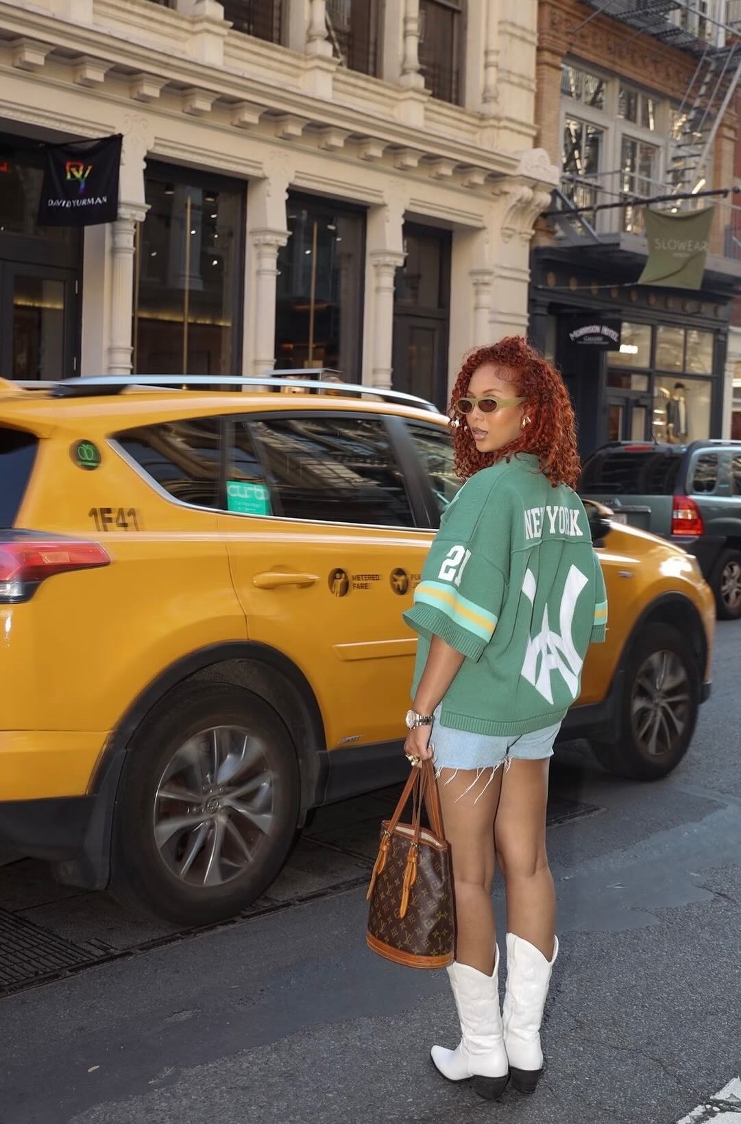 A female model standing in front of a taxi wearing a green knit jersey with New York and Wisdom Of Age logo embroidered on the back in white. 